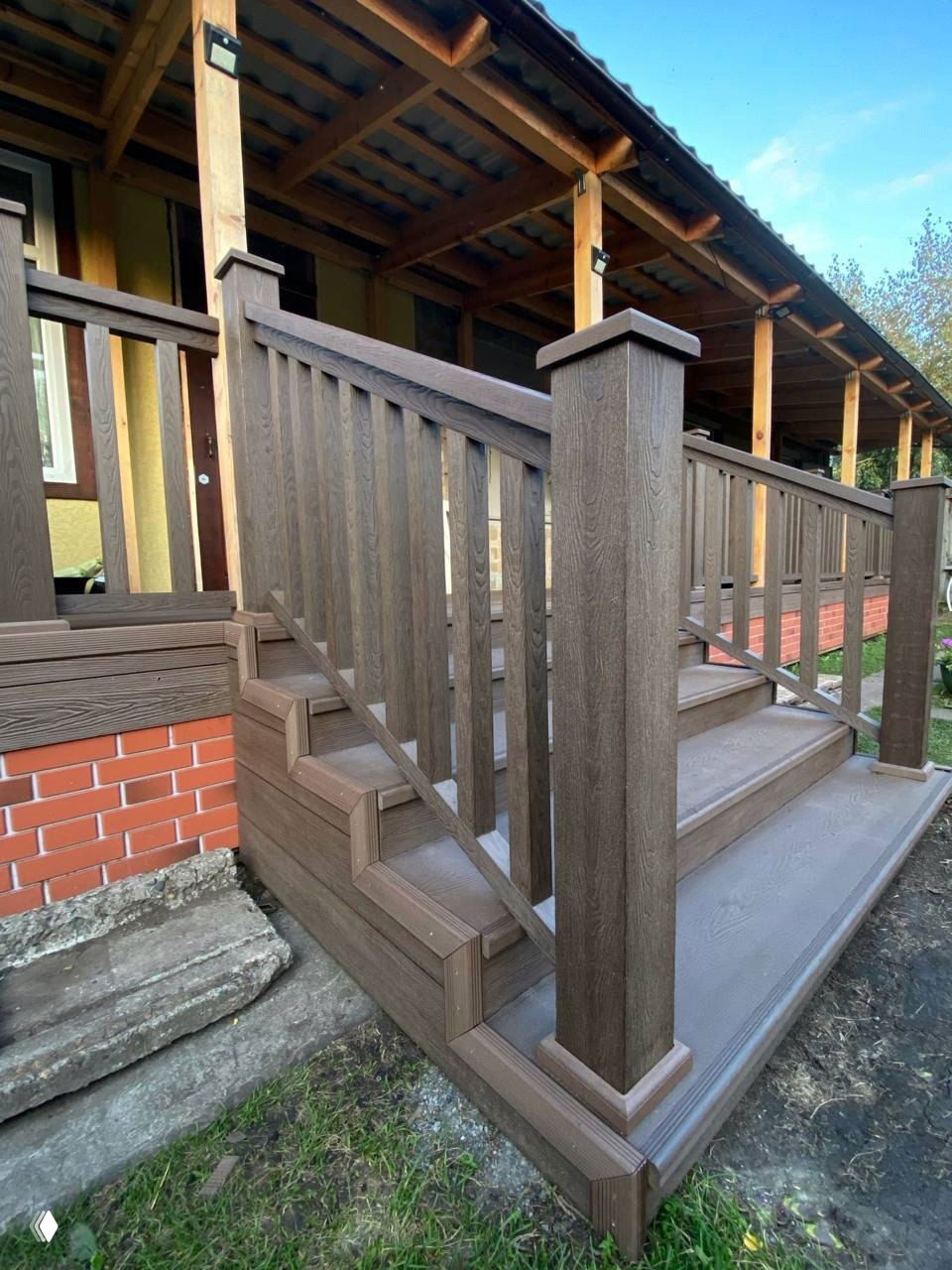 Крыльцо с аккуратными ступенями и столбами ограждения из ДПК тёмно‑коричневого цвета, декоративная отделка у входа в дом.