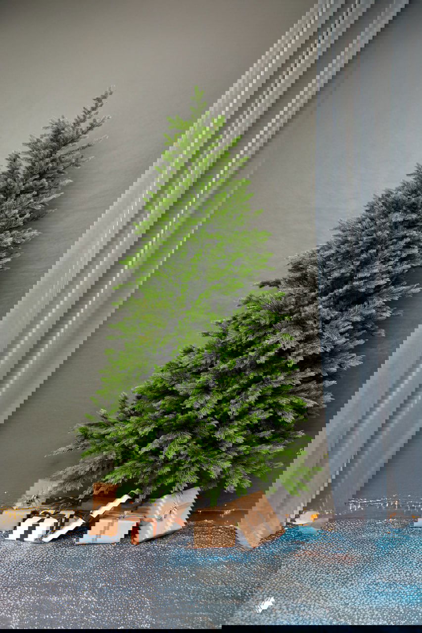 Постановочная фотография искусственной ели 1,8 м на фоне нейтральной стены с подарками у основания — вид товара для продажи.