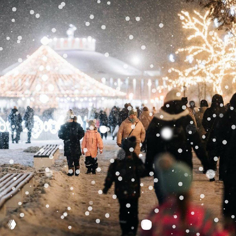 Посетители зимнего праздника в Рязани: пешеходы на заснеженной аллее перед каруселью и ярким праздничным освещением.