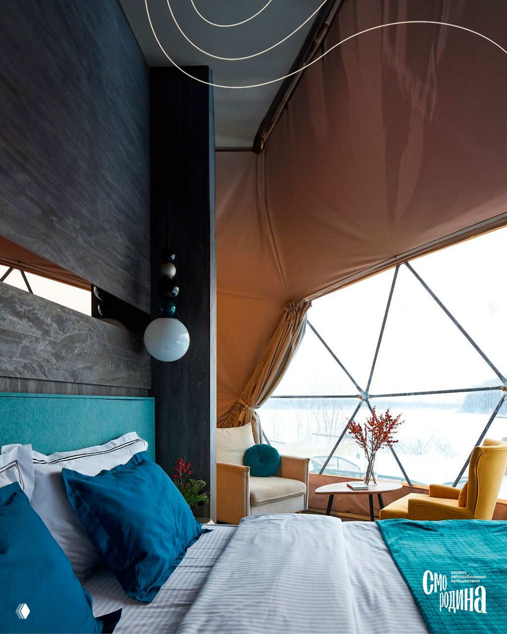 Интерьер домика: большая кровать с яркими подушками, кресла и столик у стеклянного окна, вид на снежный пейзаж за окном.