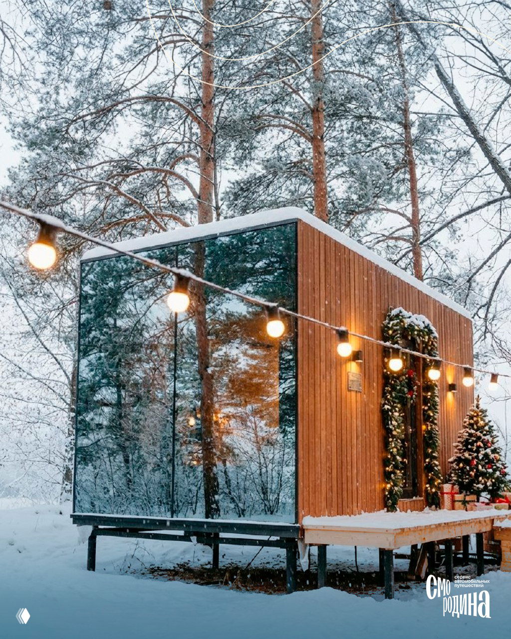 Зеркальный небольшой домик на сваях среди елей: отражения леса в стеклах, гирлянды и рождественское украшение фасада на снегу.