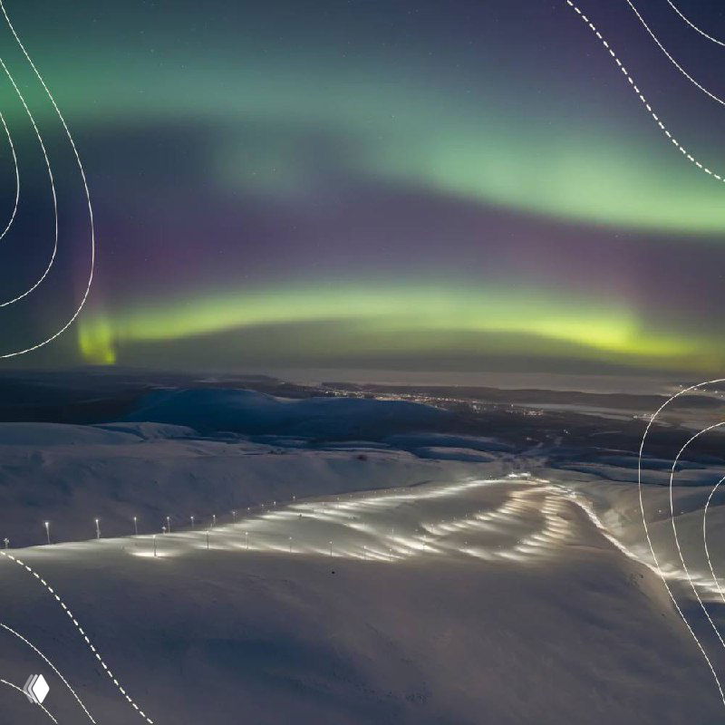 Ночное небо с зелёными и фиолетовыми переливами северного сияния над заснеженными хребтами и огнями долины вдалеке.