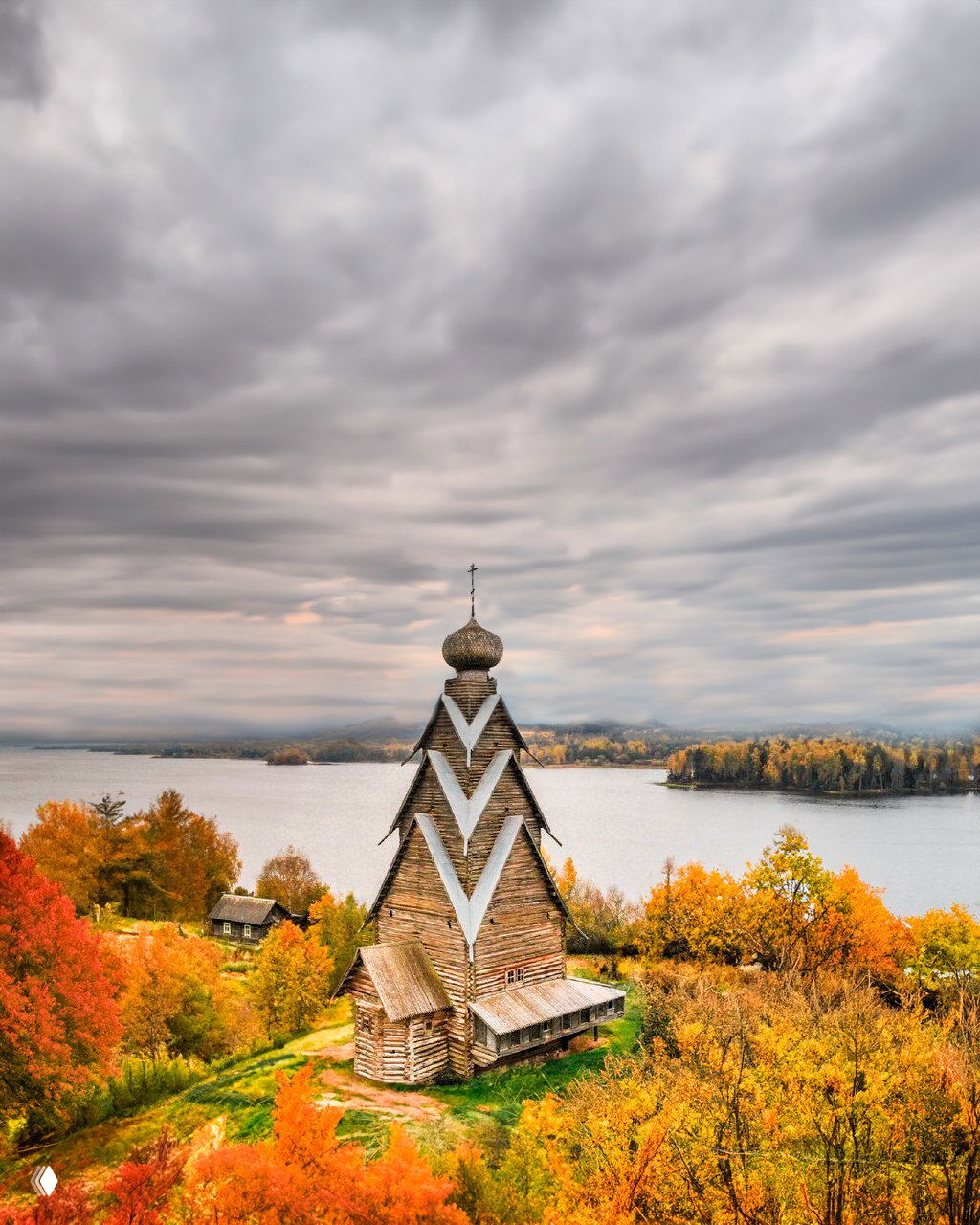 Общий план церкви Рождества Иоанна Предтечи на холме у озера Вселуг осенью: яркие осенние деревья, вода и пасмурное небо создают живописный контекст.