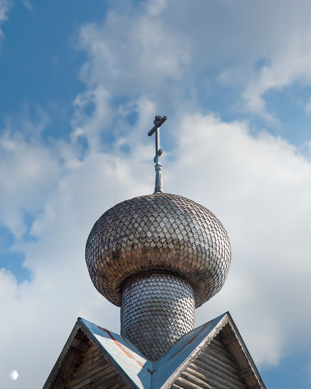 Крупный план луковичной главы и креста деревянной церкви: видна деревянная текстура, металлическая кровля и голубое небо с облаками за спиной.