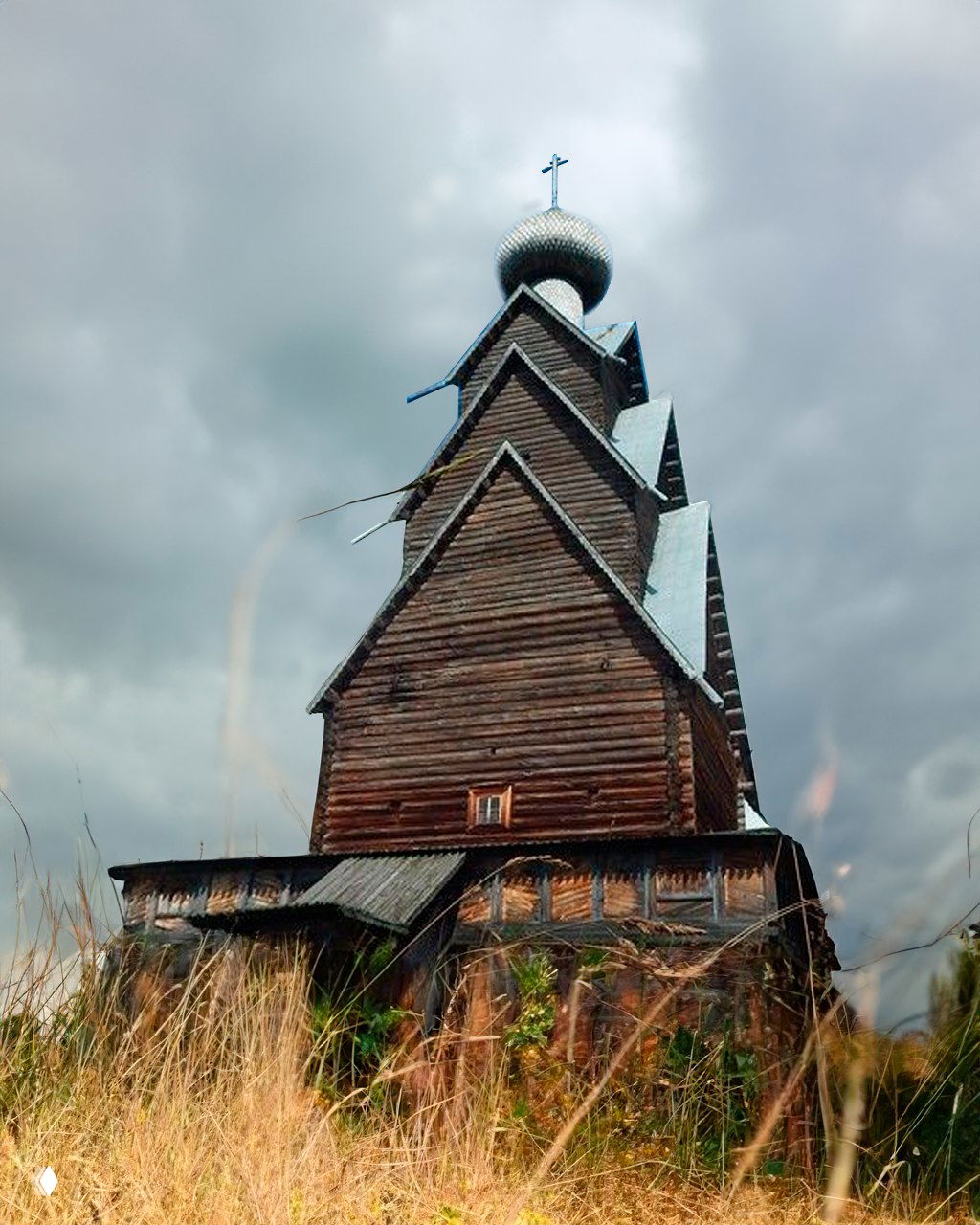 Низкий ракурс на многоярусную деревянную церковь: резные детали, трава на переднем плане и тяжёлые облака над стройной конструкцией.
