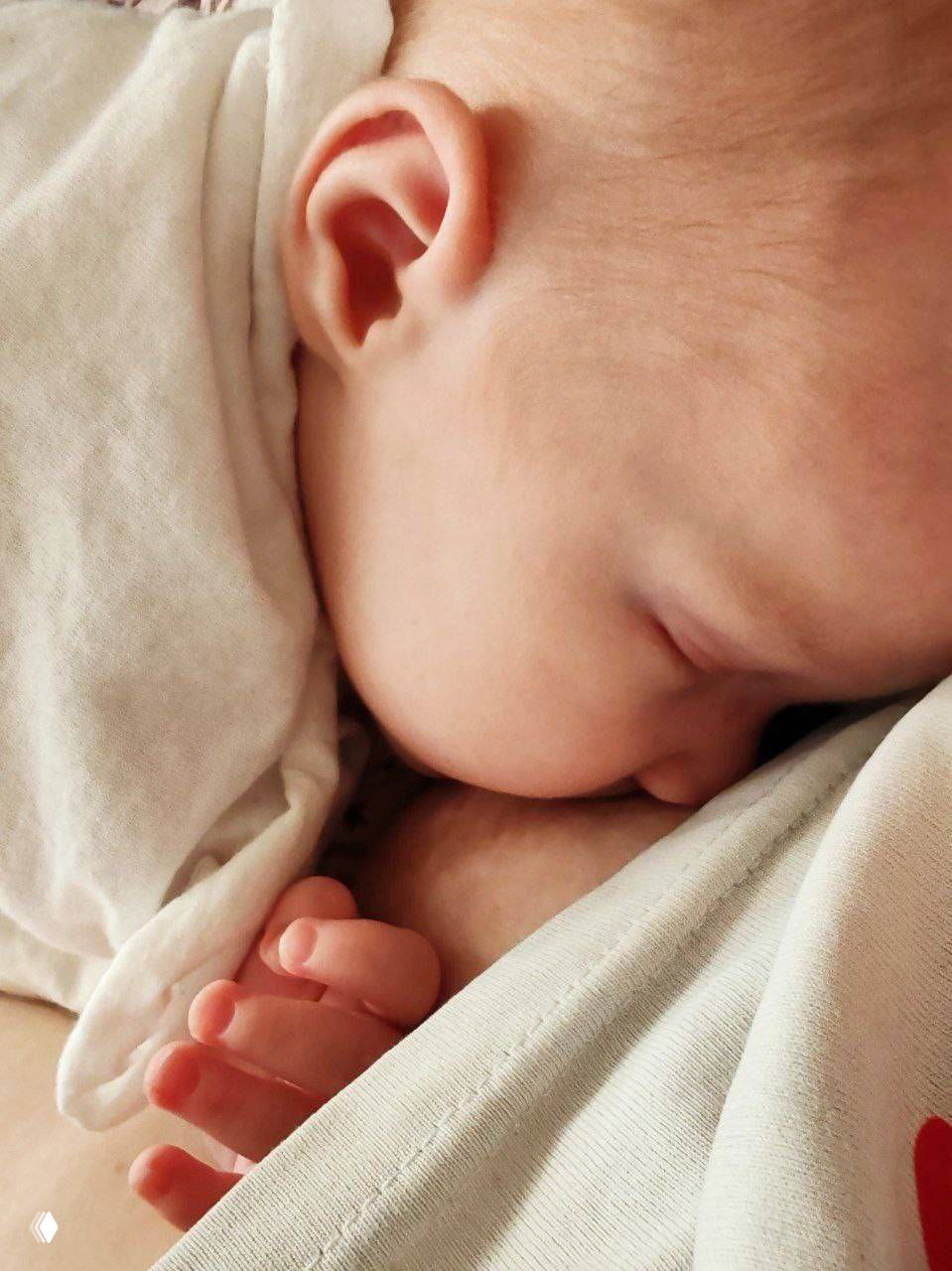 Крупный план грудного вскармливания: лицо и ухо ребёнка прижаты к груди, видны пальчики и ткань одежды, интимная домашняя сцена кормления.