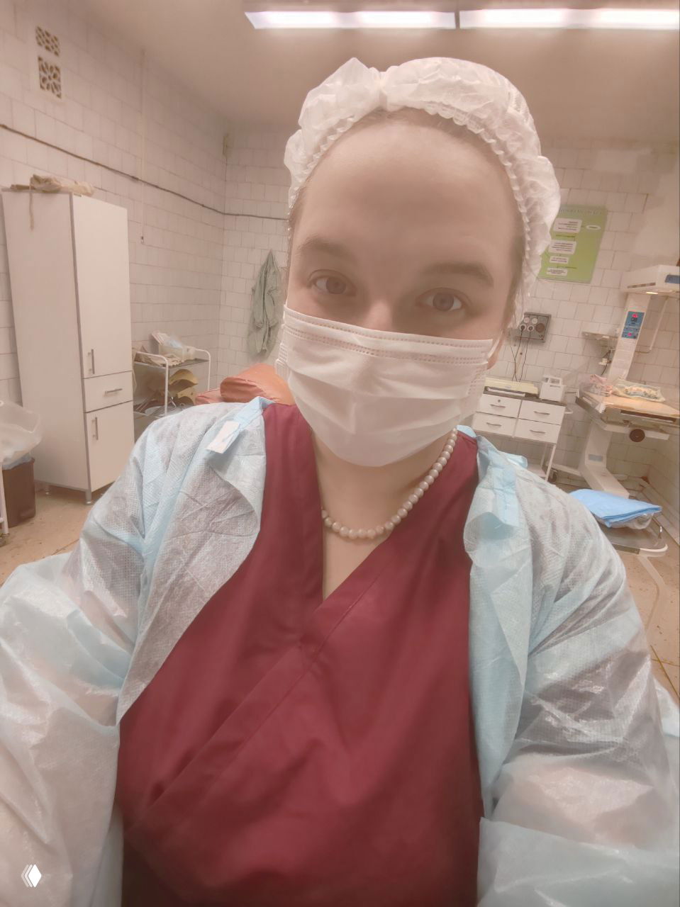 Доула в медицинской маске и одноразовой шапочке, в защитном халате, селфи в родильном зале на фоне медицинского оборудования и мебели.