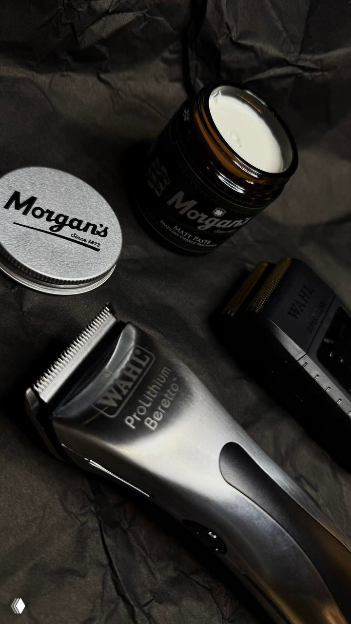 Фото под углом: металлическая машинка Wahl крупным планом рядом с баночкой пасты Morgans и насадками на тёмной фактурной поверхности.