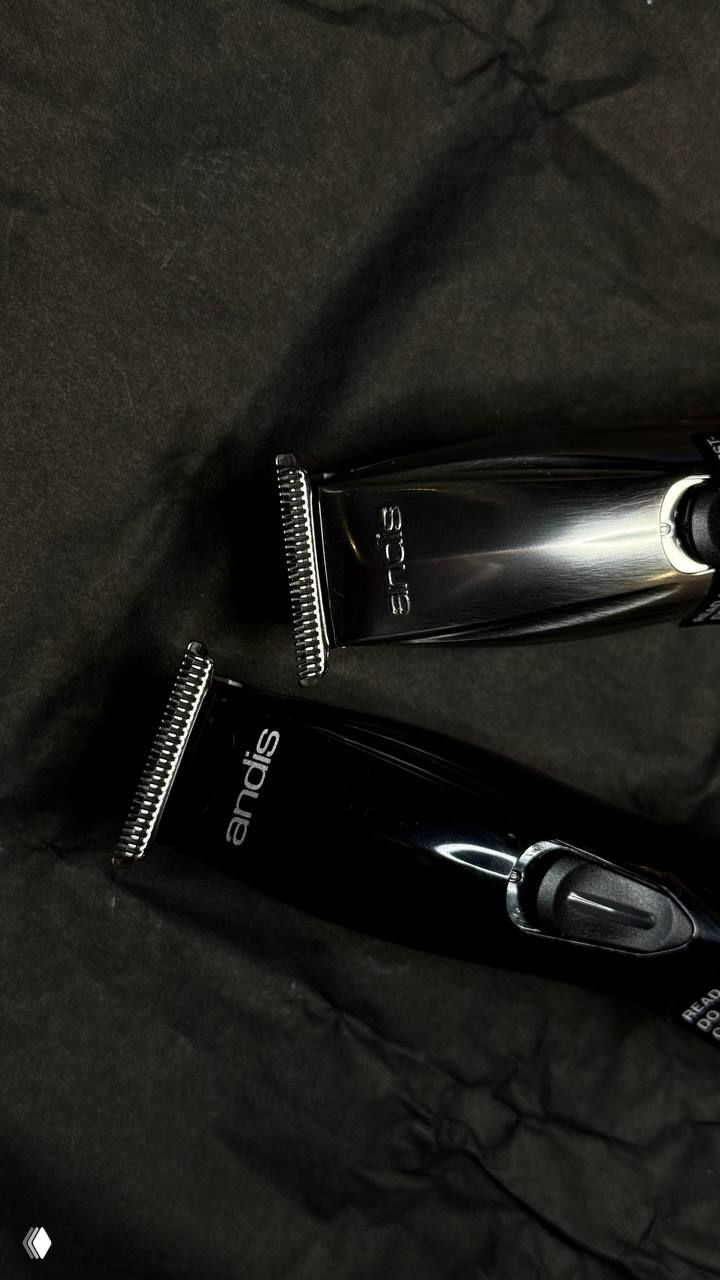 Два триммера (Andis и другое обозначение) на чёрной ткани, подчёркивающей металл и форму инструментов для стрижки и детализации.