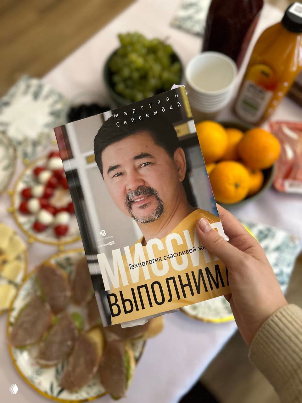Рука держит книгу «Миссия выполнима» Маргулана Сейсембая на фоне сервированного стола с фруктами, закусками и напитками — снимок с бизнес-завтрака.