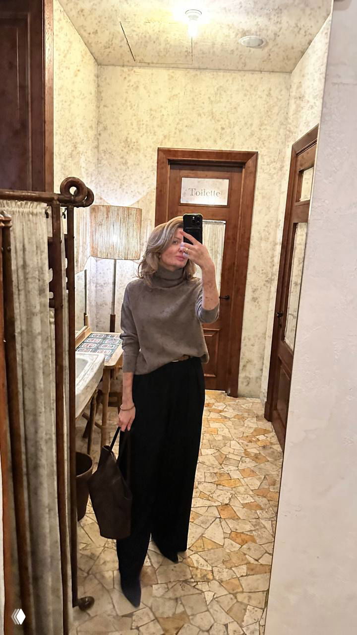 Женщина делает зеркальное селфи в коридоре у двери с табличкой «Toilette», видно интерьер с плиткой и настенной лампой.