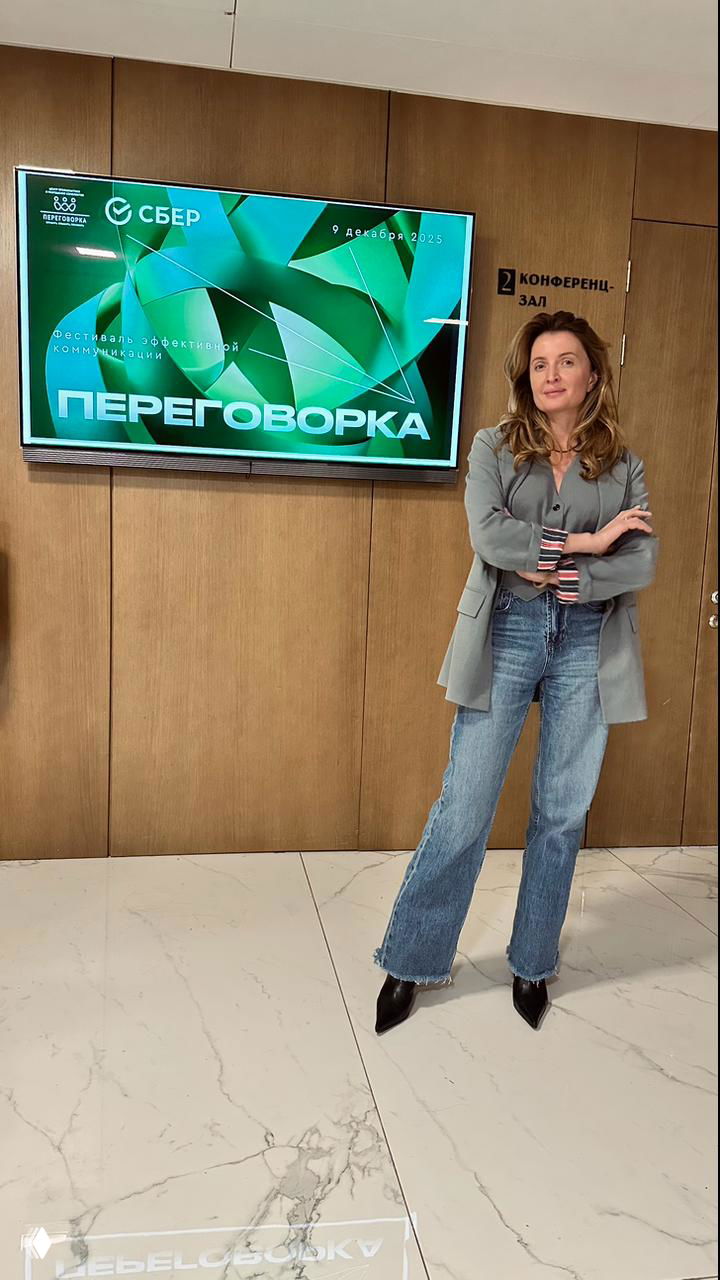 Женщина-ведущая стоит в конференц-зале рядом с экраном, на котором крупно написано «ПЕРЕГОВОРКА», виден логотип Сбер.