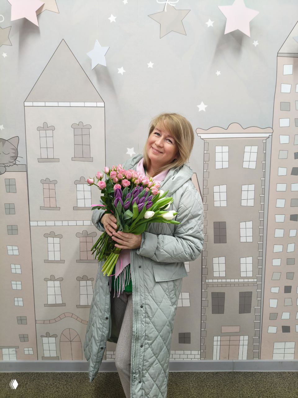Женщина держит большой букет тюльпанов на фоне декоративной стены с рисунком домов и звёзд; повседневный портрет в тёплых тонах.