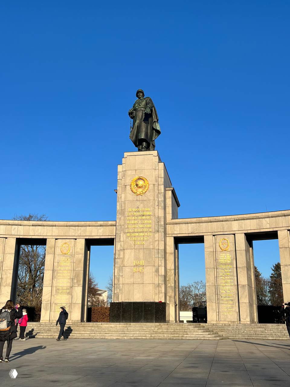 Монумент в честь советских солдат в Берлине: высокий постамент со скульптурной фигурой и золотыми надписями на фоне ясного неба.