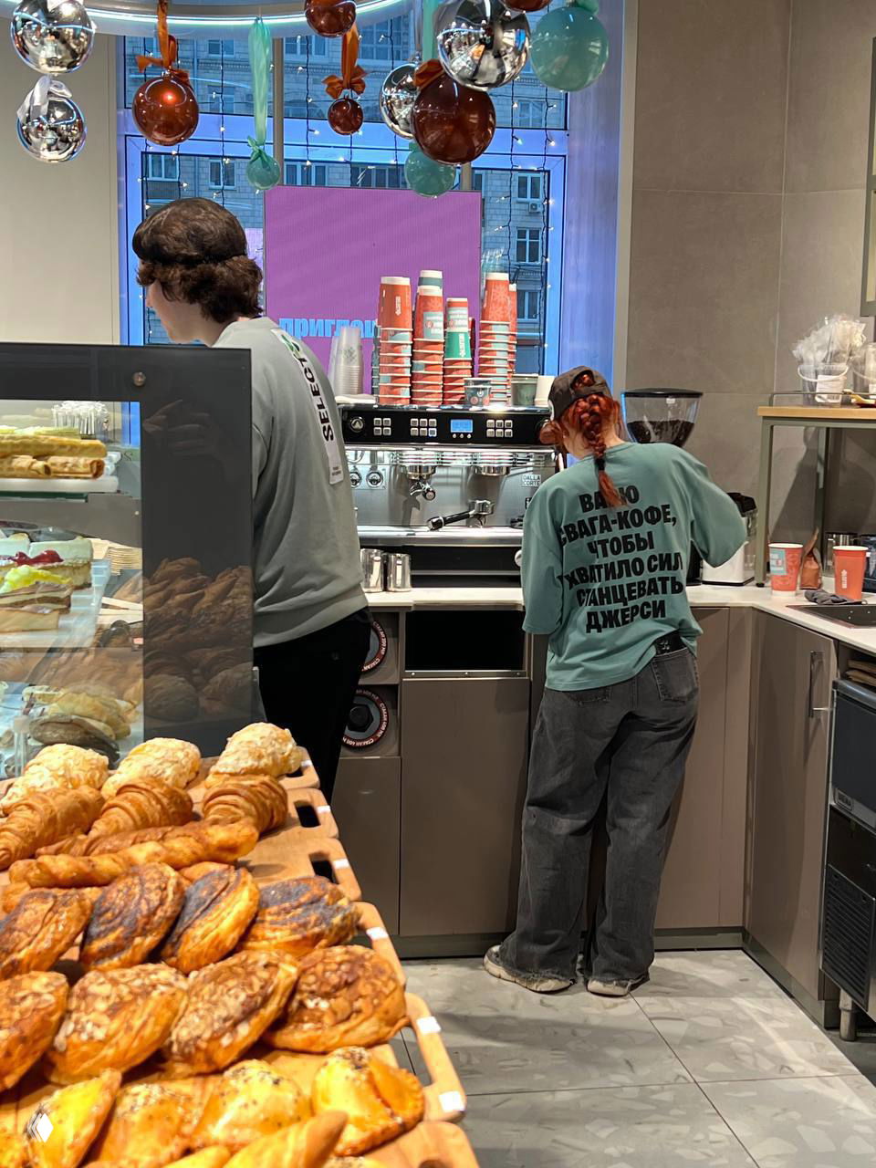 Кофейная зона и прилавок с выпечкой в Select: двое сотрудников в фирменных толстовках с надписями на спинах, передний план заполнен круассанами и булочками.