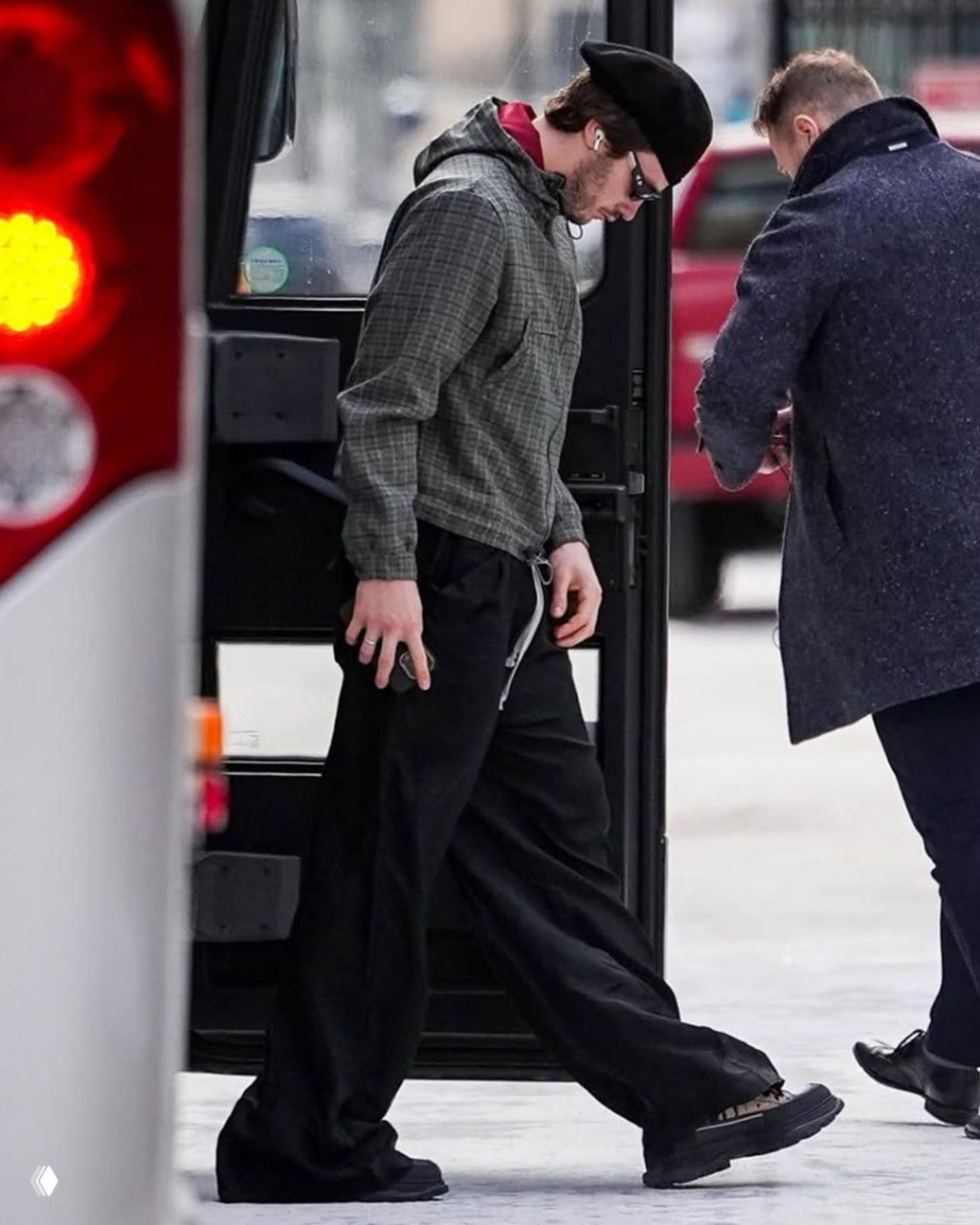 Человек в тёплой клетчатой худи, широких чёрных брюках и с наушниками выходит к автобусу на улице — уличный образ в повседневной обстановке.