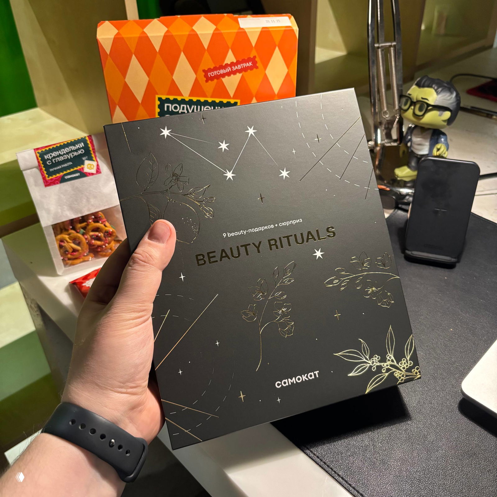 Рука держит чёрную коробку Beauty Rituals от Самоката на фоне упаковок с закусками и напитком — подарочный бьюти адвент в руке.