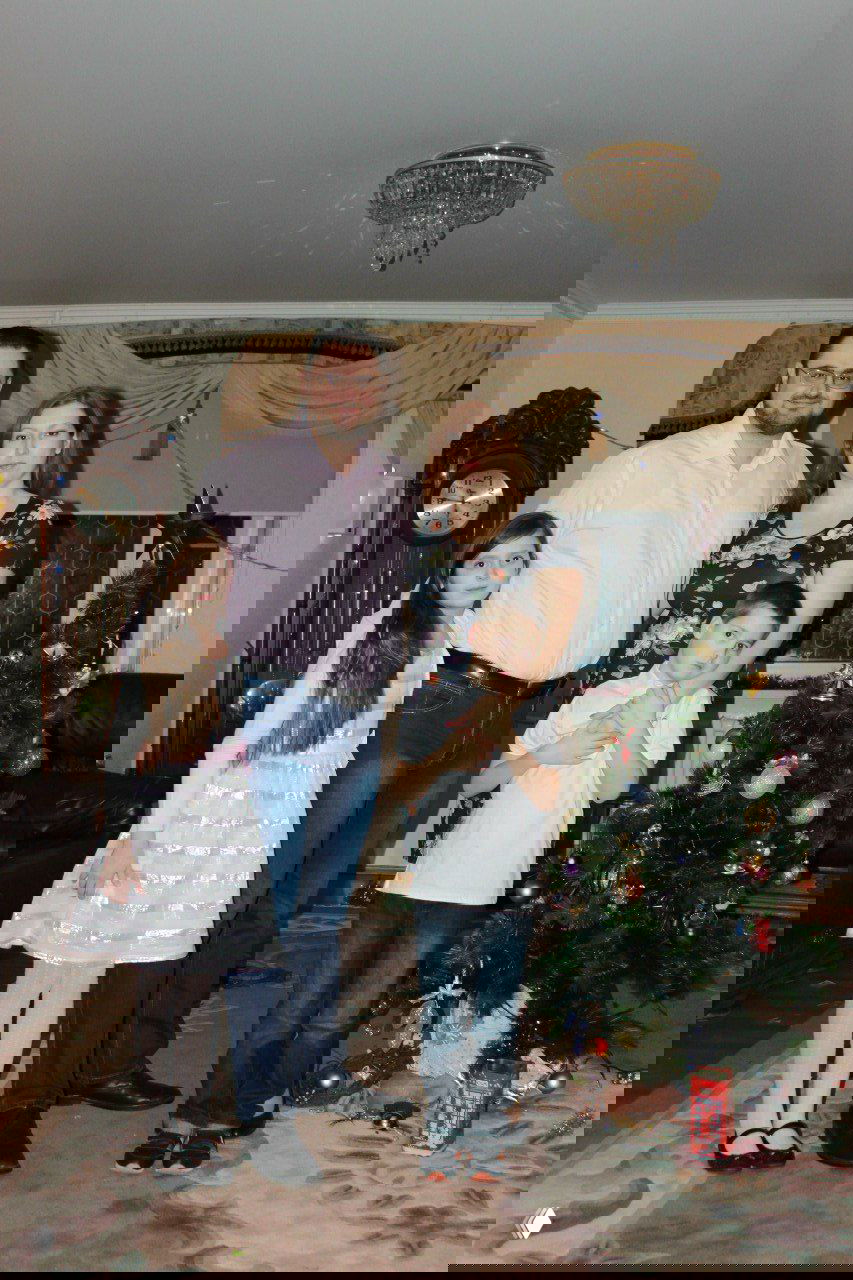 Портрет семьи у небольшой новогодней елки в гостиной: родители и двое детей стоят рядом, праздничный интерьер и часы на стене.