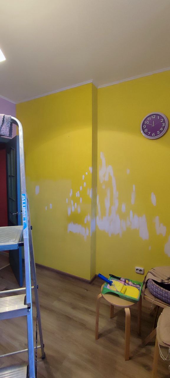 Жёлтая стена с проплешинами от шпаклёвки и фоновыми предметами: часы на стене, стремянка, табурет и элементы ремонта в комнате.