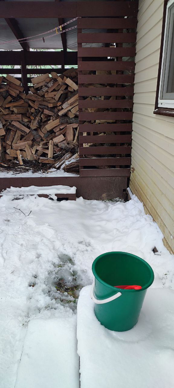Зелёное пластиковое ведро на засненном пороге рядом с кучей дров; следы воды на снегу и бытовой уголок дачи.
