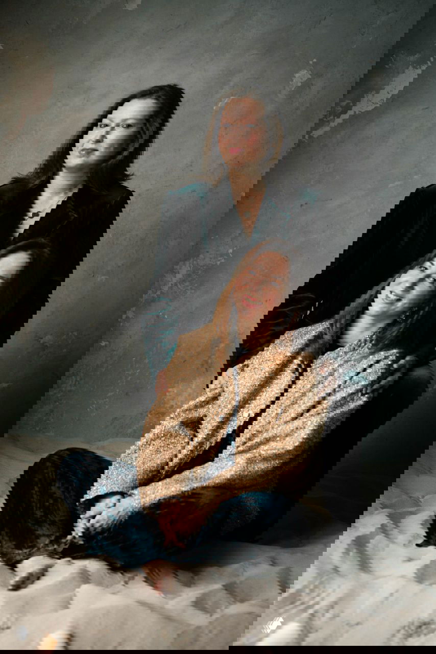 Средний план: женщина в тёмном бархатном костюме стоит за подругой в блестящем пиджаке на фоне фактурной стены — тёплая студийная эстетика.