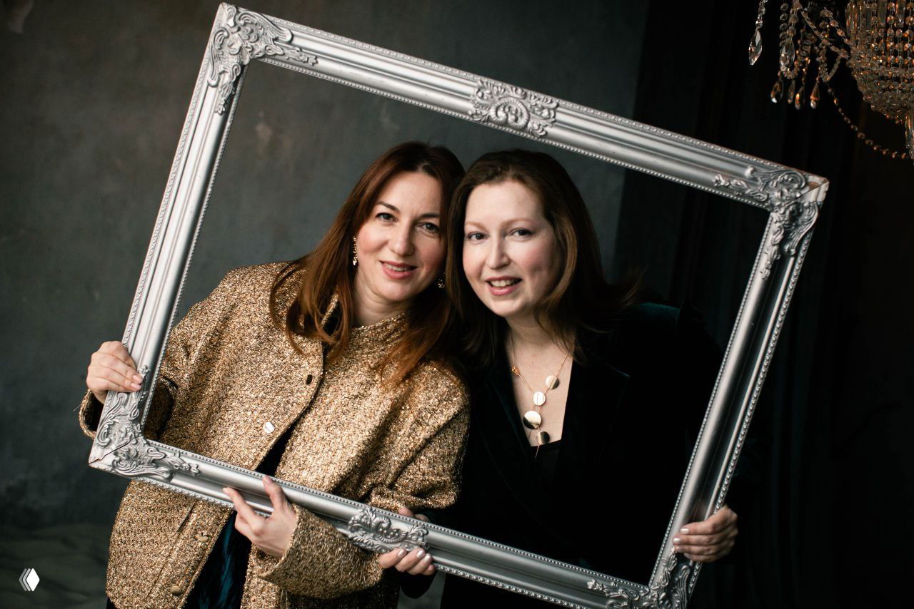 Портрет двух женщин за деревянным столом: одна обнимает другую за плечи, тёплый домашний антураж и ощущение поддержки.