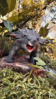 Фотография фигуры динозавра среди тропической зелени в парке Gulong; ракурс подчёркивает масштаб и эффект присутствия.