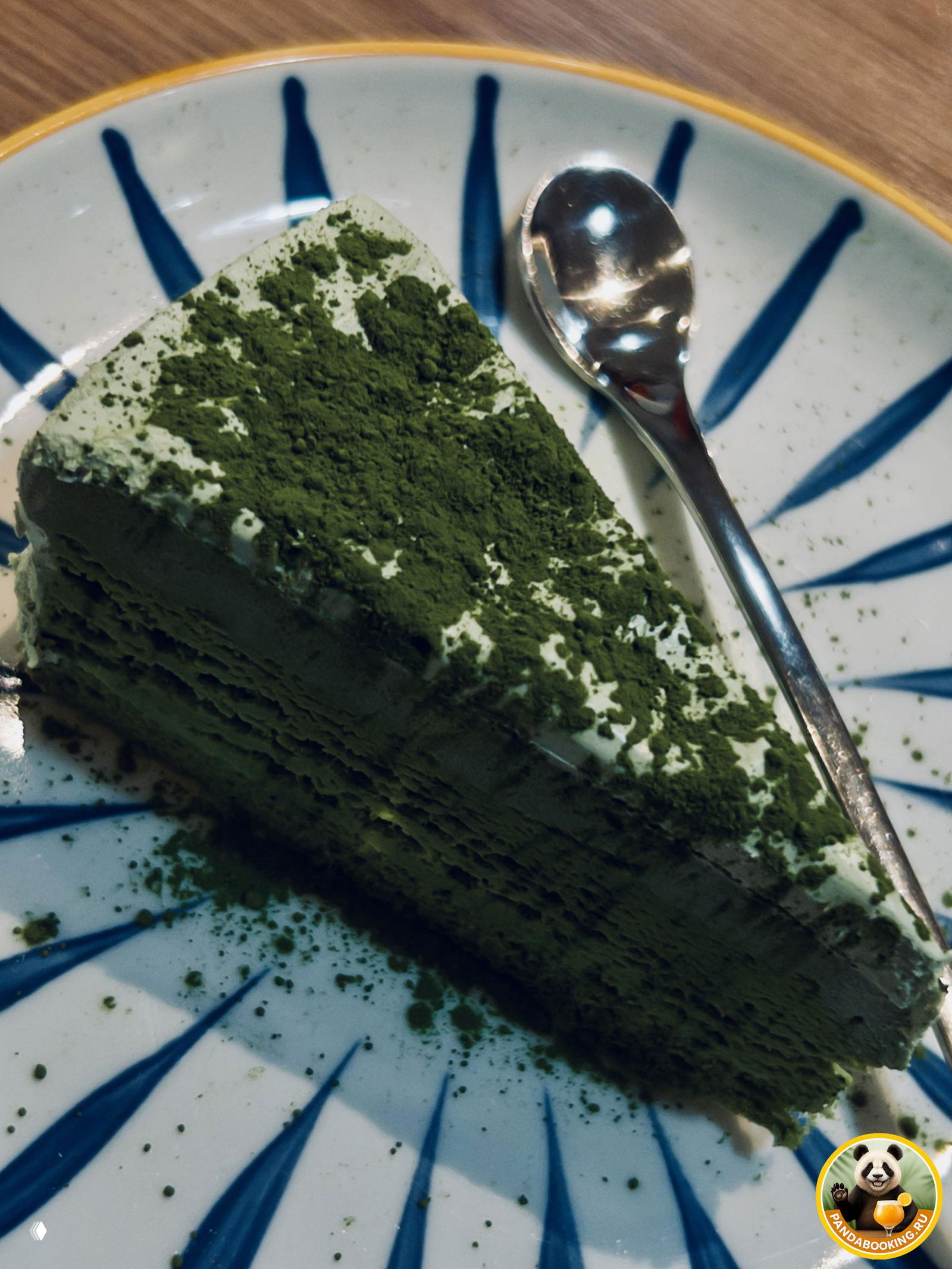 Кусок десерта на белой тарелке, похоже на торт с зелёным порошком матча сверху и ложкой рядом, подан в кафе рядом с основным блюдом.