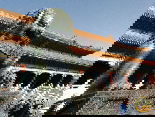 Бронзовый китайский лев-страж у входа в дворцовый комплекс, мощная скульптура на каменном постаменте на фоне традиционной кровли.