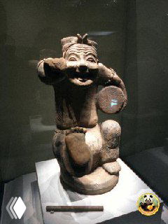 Керамическая музейная фигурка-артефакт в витрине, забавная поза и мягкое музейное освещение подчёркивают историческую ценность предмета.