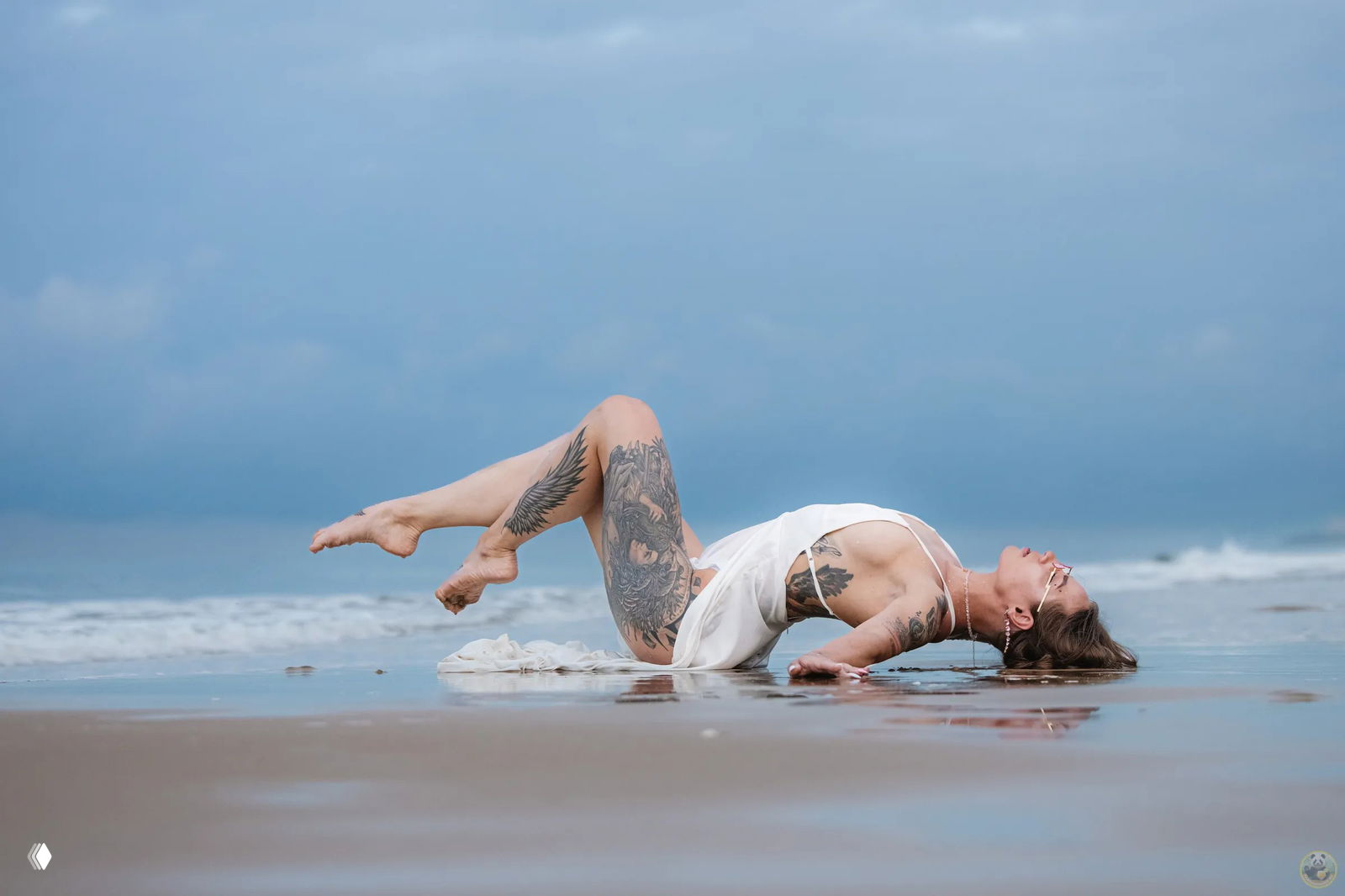 Женская пляжная фотосессия на мокром песке в Санье: модель в белом платье с большими татуировками лежит на берегу, океан и облачное небо на заднем плане.