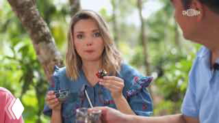 Женщина в тропическом саду пробует угощение во время съёмки телепередачи о Хайнане; видны зелёные растения, стол и микрофон на переднем плане.