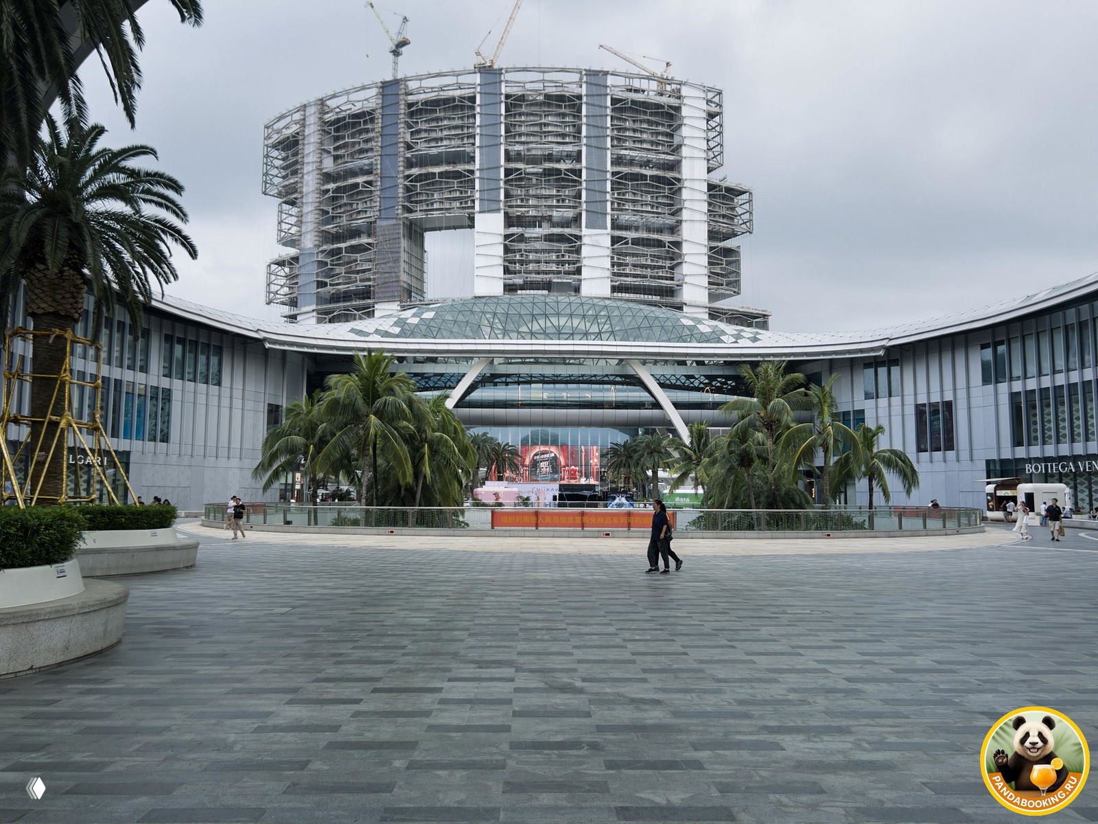 Внешний фасад крупного торгового комплекса Duty Free в бухте Хайтанвань: широкая площадь, пальмы и арочная входная часть на фоне пасмурного неба.