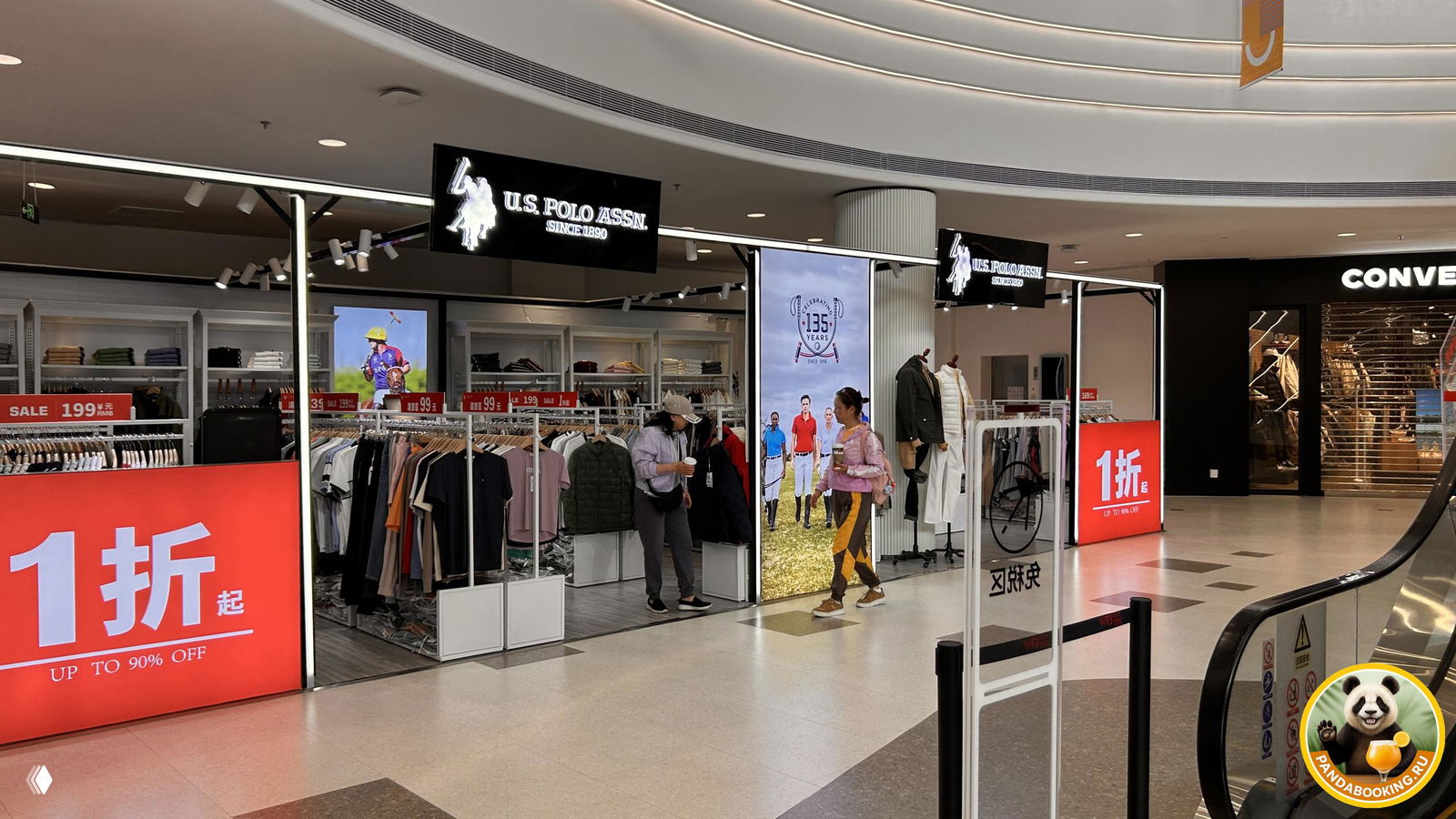 Небольшие стенды и павильоны с одеждой и вывеской U.S. Polo Assn., цифровые табло с акциями и движением посетителей у эскалатора.