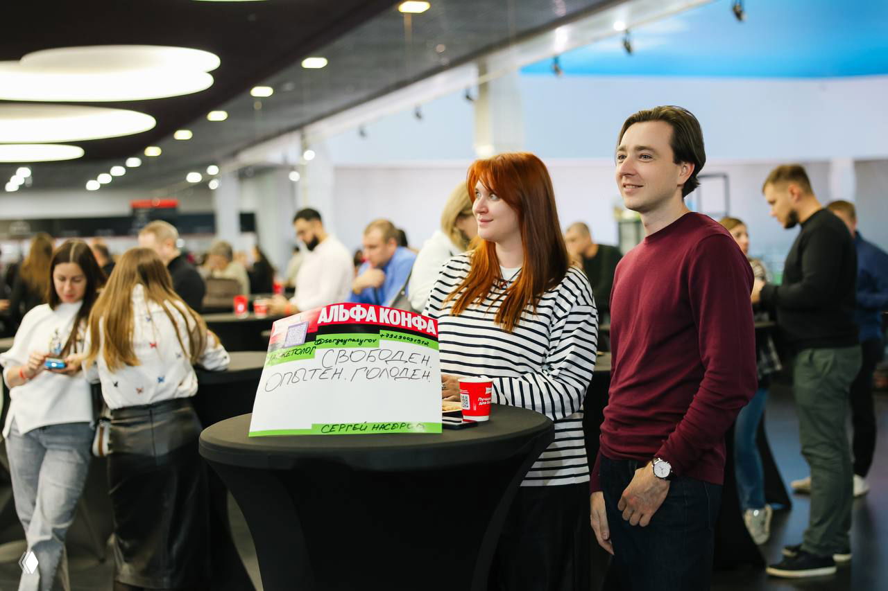 Участники в лобби конференции у круглого стола; на столе плакат с надписью «Свободен. Опытен. Голоден», люди рядом улыбаются и общаются.