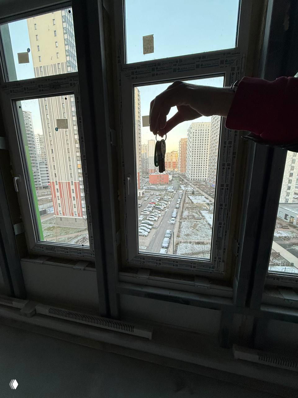Рука держит ключи у окна, вид на городской двор и парковку зимой — символ передачи ключей от будущего офиса и начало ремонта.