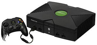 Фотография оригинальной консоли Xbox с контроллером на белом фоне: вид спереди, ретро‑дизайн, зелёный логотип и кабели, подчёркивающие эпоху 2000-х.