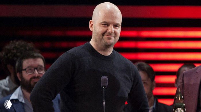 Человек в тёмной футболке у микрофона на сцене; на заднем плане — красные световые полосы, в момент награждения или выступления.