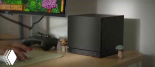 Кубическая консоль Steam Machine на рабочем столе рядом с монитором и контроллером — компактный тёмный корпус, вид в интерьерной обстановке