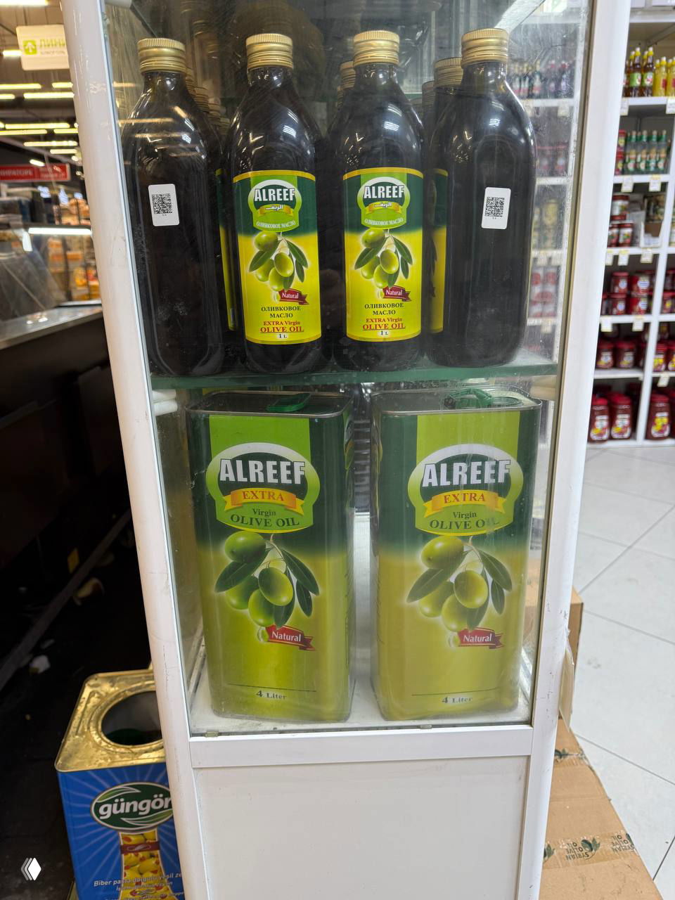 Жестяные банки оливкового масла ALREEF 4 л и бутылки на полке холодильника в сирийском магазине, хорошо видны этикетки и упаковка.