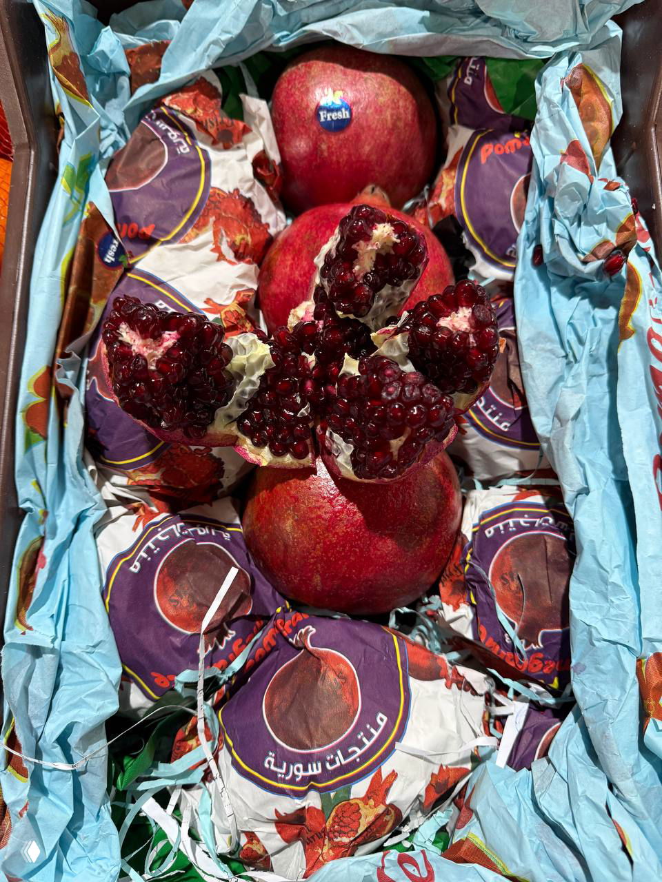 Коробка сирийских гранатов: разрезанный плод с крупными густо-бордовыми зернами на фоне фирменных бумажных обёрток и упаковки.