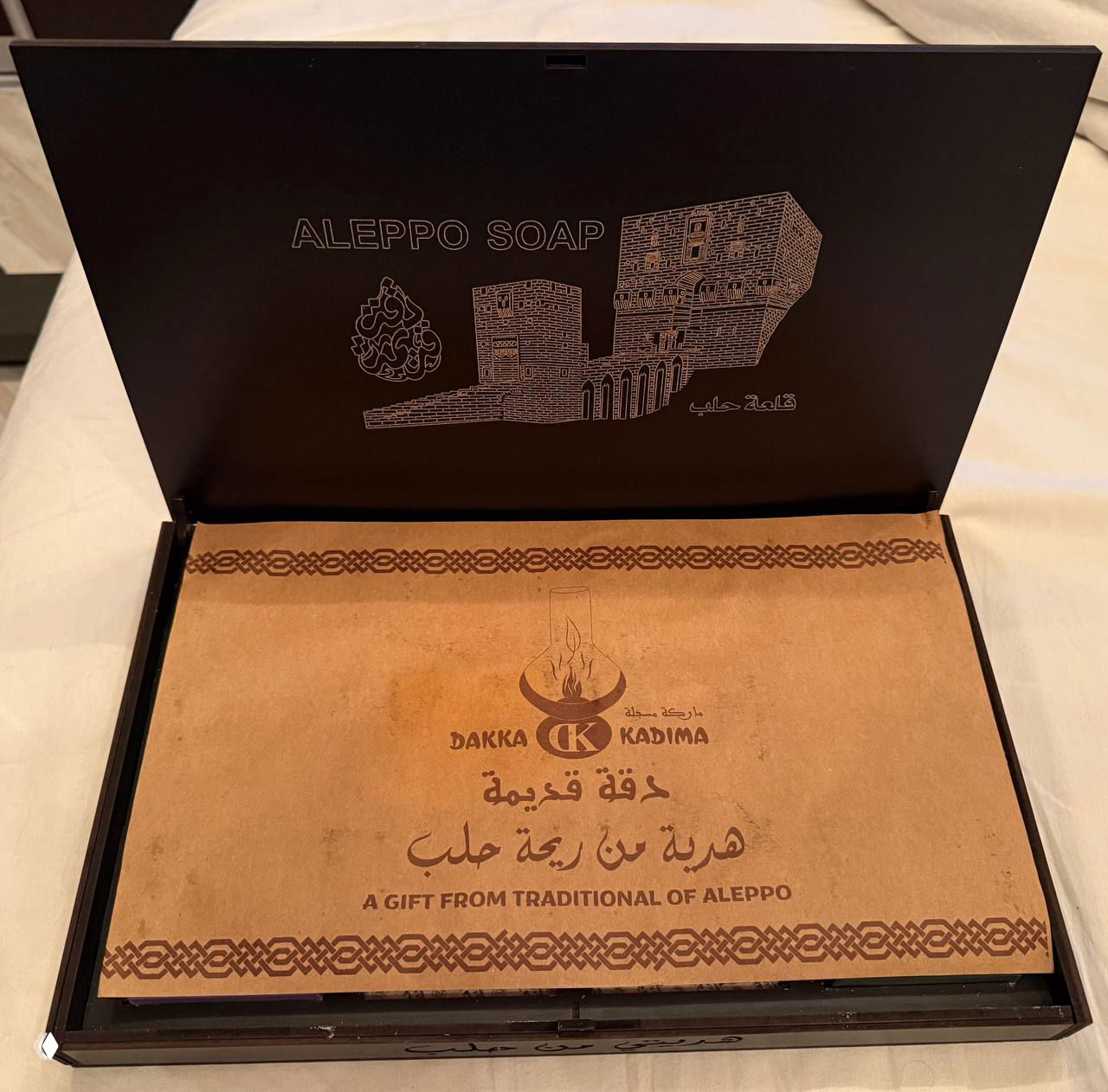 Открытая коробка с бумажной вставкой и печатью «ALEPPO SOAP», видна декоративная полиграфия и арабский логотип внутри крышки.