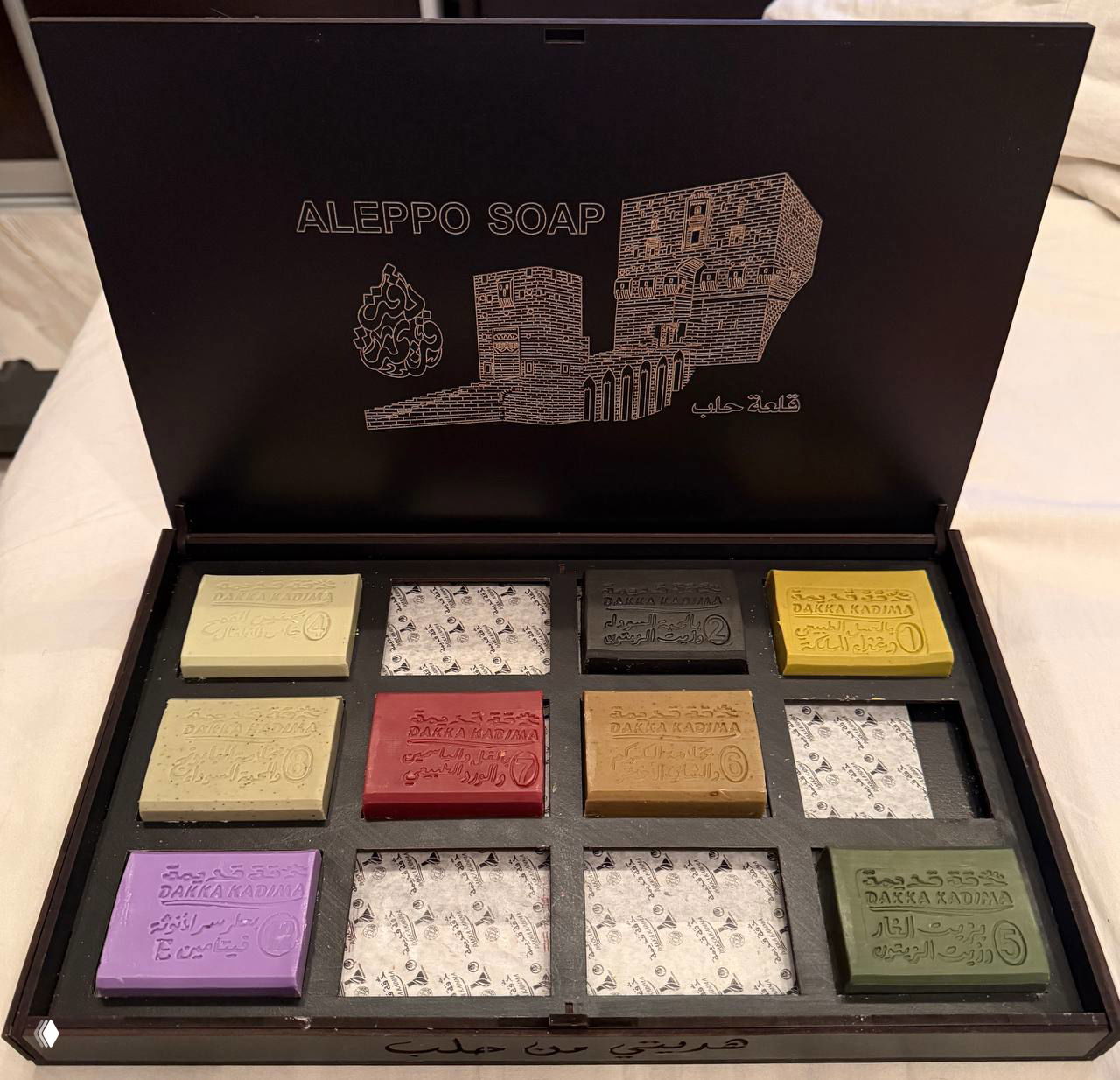 Набор кусочков алеппского мыла разных оттенков, уложенных в секционированную коробку — аккуратно расположенные прямоугольные бруски.