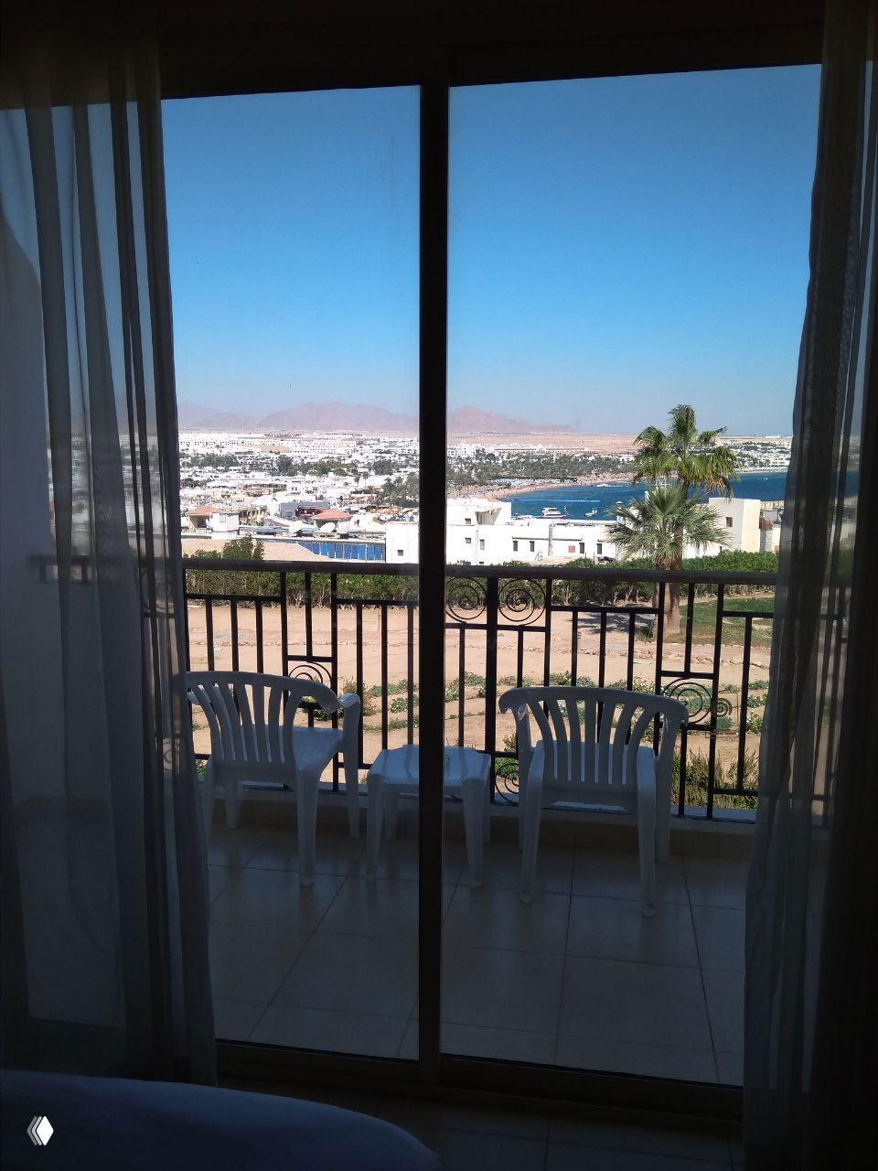 Вид из комнаты через раздвижное стекло на балкон с двумя пластиковыми стульями, дальше город, пальма и бухта на горизонте — тихая сцена для отдыха.
