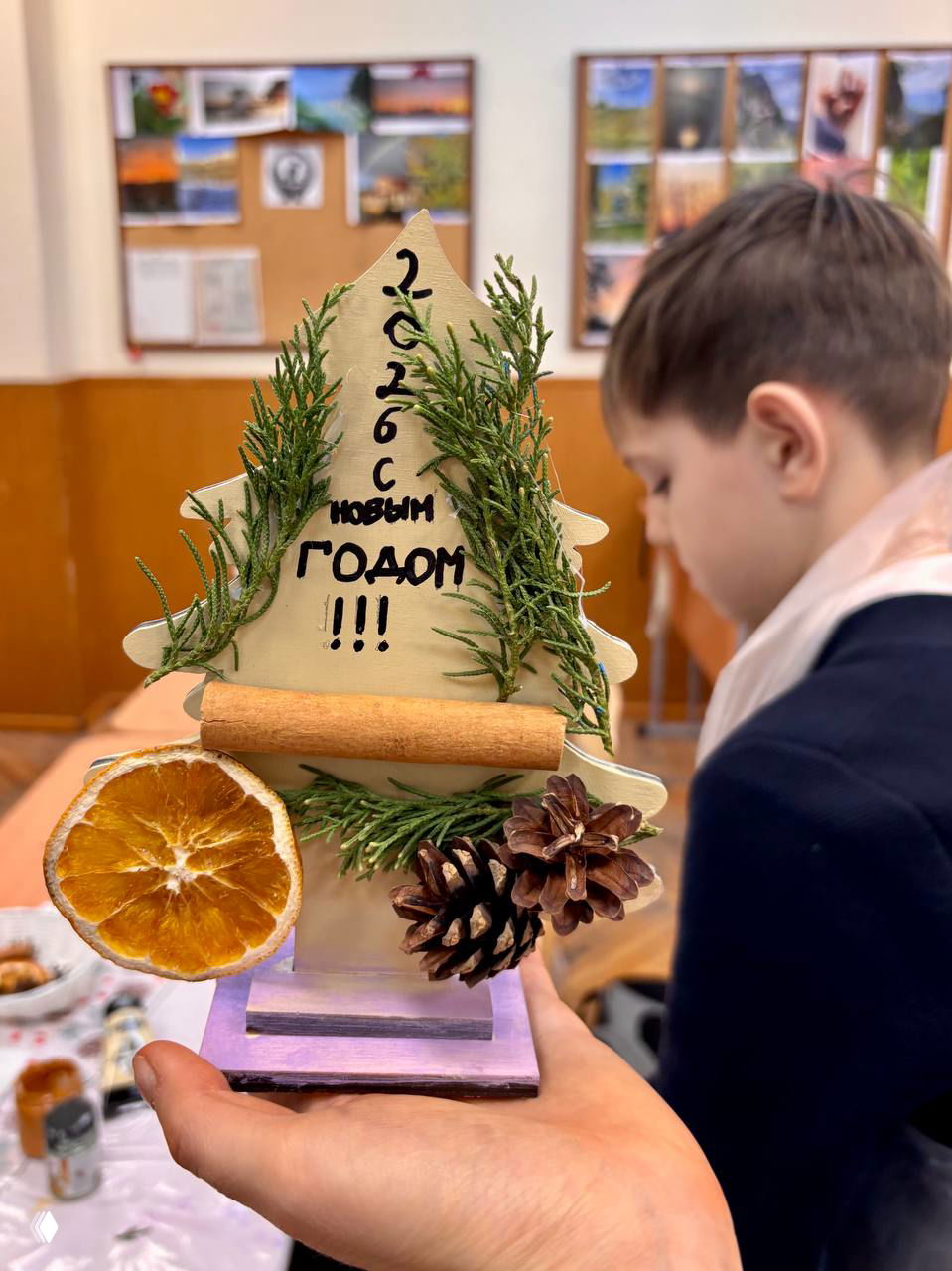 Ближайший план деревянной ёлочки с надписью «2026 с Новым годом!!!» и элементами декора; на заднем плане школьник.
