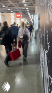 Коридор выставочного павильона с посетителями, идущими между стендами; видно людей с сумками и общую активность мероприятия.