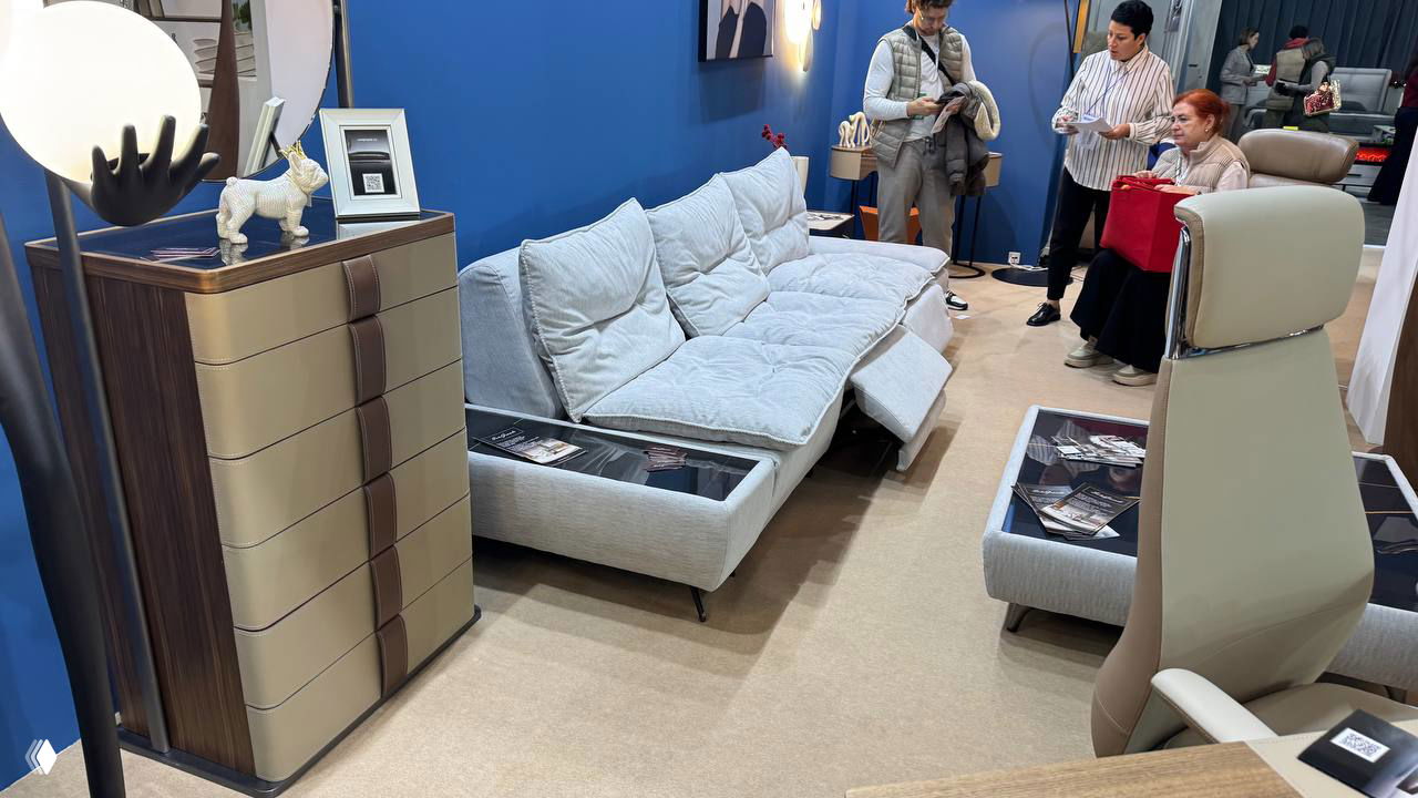 Композиция дивана и комодов у синей стенки стенда; мягкая мебель в нейтральных тонах и посетители, рассматривающие образцы.