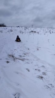 Человек на ватрушке спускается по снежному холму; заснеженный склон, следы и серое небо создают атмосферу активного зимнего отдыха и перезагрузки.