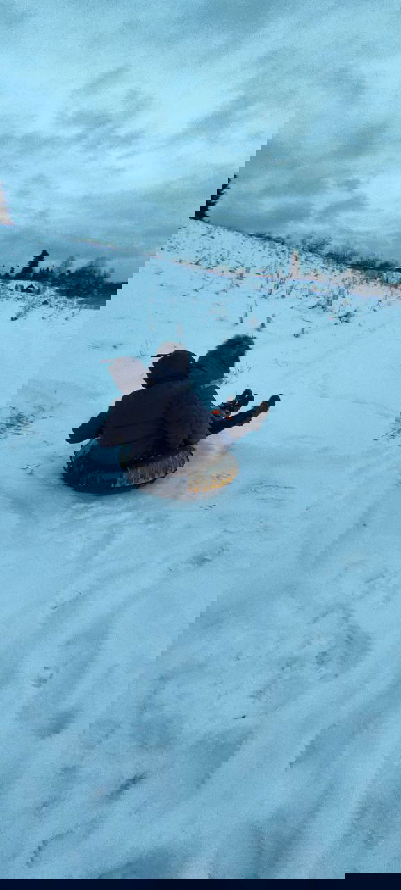 Двое катаются на надувной ватрушке по снежному склону; следы и деревенский пейзаж вдали под хмурым зимним небом, ощущение простого удовольствия.