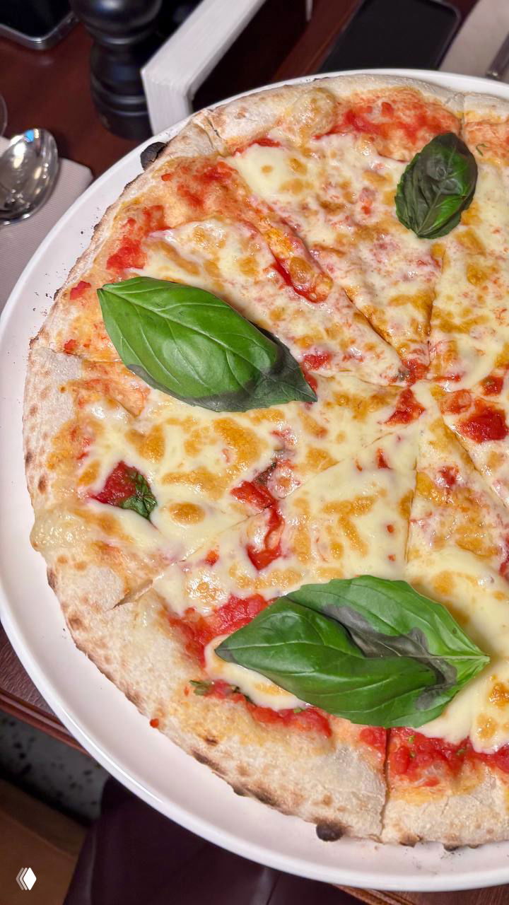 Пицца Маргарита на белой тарелке: тонкая корочка, томатный соус, расплавленный сыр и листья базилика сверху.